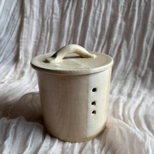 Könnte beinhalten: Eine cremefarbene Butterdose aus Keramik mit Deckel und Griff. Der zylindrische Behälter hat drei kleine Löcher an der Seite. Der Deckel hat einen gebogenen Griff. Die Dose steht vor einem weichen, strukturierten Hintergrund.