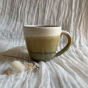 May include: A handmade ceramic mug with a rounded body and a curved handle. The mug features a gradient of colors, starting with a cream-colored rim, transitioning to a golden-yellow middle, and ending with a green base. The mug is displayed on a white, textured surface.