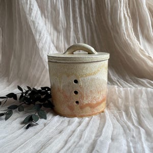 May include: A ceramic container with a lid and handle. The cylindrical body has a gradient of colours, transitioning from beige to a warm peach hue. Three small, evenly spaced holes are visible on the side. The lid has a curved handle.