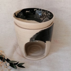 Könnte beinhalten: Ein weißer Keramik-Öldiffusor mit einem schwarzen abstrakten Design. Der Diffusor hat eine abnehmbare obere Schale für ätherische Öle und einen unteren Abschnitt mit einer Öffnung für ein Teelicht.