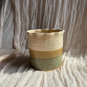 May include: A handcrafted ceramic vase with a cylindrical form and a wavy edge. The vase displays horizontal bands of colour: a light beige top, a golden-yellow middle, and a muted green base. The vase rests on a neutral-coloured, textured fabric.