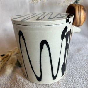 May include: A cream-coloured ceramic container with a lid, decorated with a black dripped paint design. The lid and container have a wavy black pattern. It has a small wooden handle attached to the side.