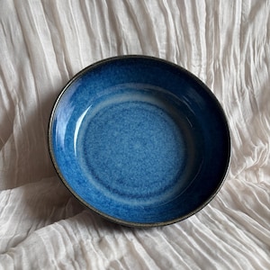 May include: A round, blue ceramic bowl with a dark rim. The bowl has a glossy finish and a textured interior. The bowl is set against a white, textured background. This bowl is suitable for serving food.