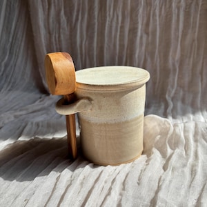 Peut inclure: Un beurrier en céramique avec un couvercle et une poignée en bois. Le plat cylindrique présente un design bicolore, avec un dessus beige clair et une base beige plus foncée. Le couvercle en bois a une poignée arrondie.