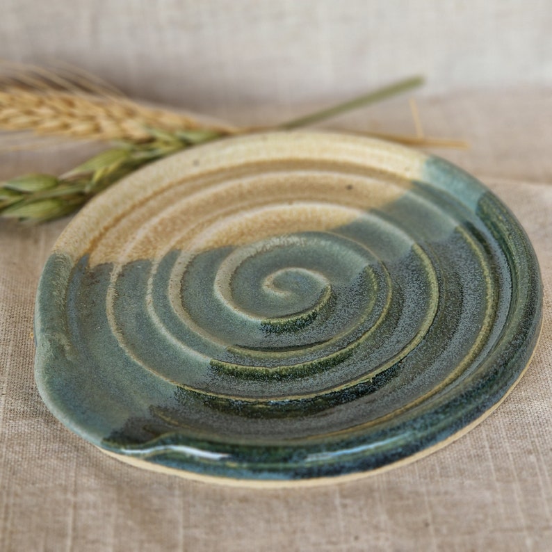 May include: A handmade ceramic spoon rest with a spiral design. The spoon rest features a blend of tan, green, and blue glazes. The circular shape has a small indentation for resting utensils. The background is a neutral linen fabric.