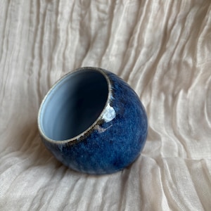 May include: A small, round ceramic bowl with a deep blue glaze. The interior is a lighter blue, and the rim has a dark brown accent. The bowl is set against a textured, off-white fabric background. The bowl's shape is slightly tilted.