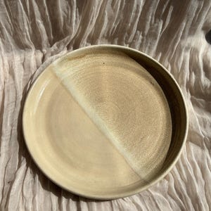 May include: A handmade ceramic plate with a two-tone design. The plate is divided diagonally, with one half a light beige and the other a darker tan. The rim is a darker brown. The plate is likely used for serving food.