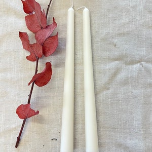 May include: Two white taper candles on a beige linen background. The candles are long and slender, with a pointed tip.