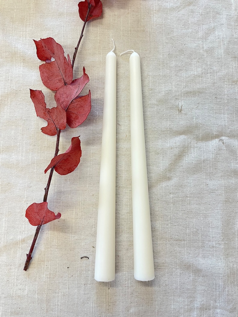 May include: Two white taper candles on a beige linen background. The candles are long and slender, with a pointed tip.