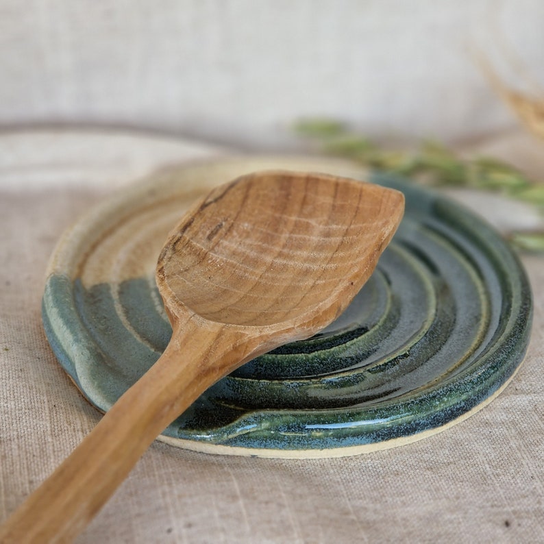 May include: A wooden spoon rests on a handmade ceramic spoon rest. The spoon rest is circular with a blue-green glaze and a beige section. The wooden spoon is light brown.