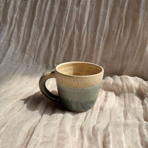 May include: A handmade ceramic mug with a rounded handle. The mug features a two-tone design, with a beige rim and a gradient of green and blue hues on the lower half. The mug is set against a neutral, textured background.