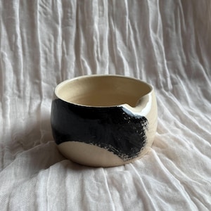 May include: A ceramic bowl with a cream-colored exterior and interior. A bold black brushstroke design wraps around the bowl's exterior. The bowl has a small spout on the rim. The bowl is sitting on a white, textured surface.