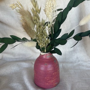 May include: A small, round, pink ceramic vase with a textured surface, holding a bouquet of dried flowers and greenery. The arrangement includes green eucalyptus leaves, beige grasses, and white fluffy accents. The vase has a white rim.