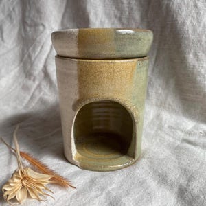 May include: A ceramic oil warmer with a two-tone design. The top dish is light green and the base is a mix of beige and green. It has an arched opening on the front. A dried flower and stem are in the foreground.