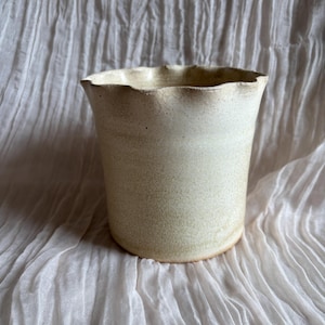 Könnte beinhalten: Handgefertigte Keramikvase mit cremefarbener Außenseite und leicht strukturierter Oberfläche. Der Rand hat ein einzigartiges, wellenförmiges Design. Die Vase ist zylindrisch und steht auf weißem Stoff.