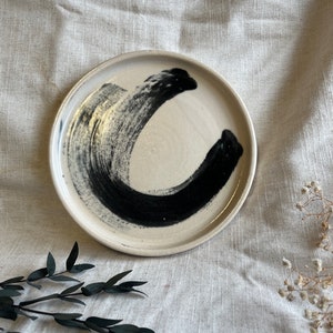 May include: A white ceramic plate with a black brushstroke design. The plate is round and has a slightly raised rim.