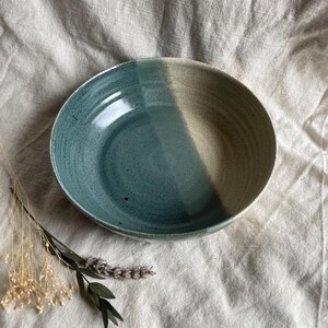 May include: A large, ceramic bowl with a light blue glaze on one side and a beige glaze on the other side. The bowl is sitting on a white fabric surface.