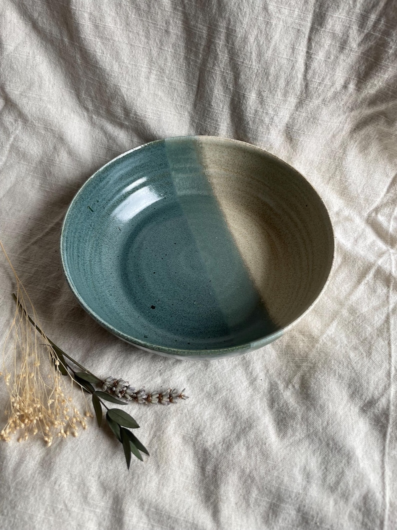 May include: A large, ceramic bowl with a light blue glaze on one side and a beige glaze on the other side. The bowl is sitting on a white fabric surface.