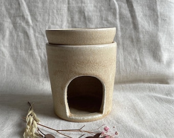 May include: A ceramic oil burner with a white glaze and a small opening in the front. The burner is made of two pieces, a top dish and a bottom base. The burner is sitting on a white fabric surface.