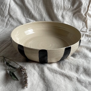 May include: A large, white ceramic bowl with black stripes around the rim. The bowl is sitting on a white fabric surface.