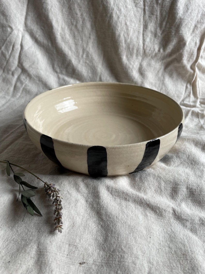 May include: A large, white ceramic bowl with black stripes around the rim. The bowl is sitting on a white fabric surface.