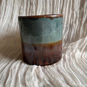 Könnte beinhalten: Ein handgefertigter Keramikbecher mit zweifarbigem Glaze. Die obere Hälfte ist ein gedämpftes Türkis, das in ein sattes, erdiges Braun übergeht. Der Rand hat einen dunkelbraunen Akzent. Der Becher steht auf einer weißen, strukturierten Oberfläche.