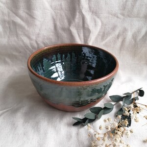 May include: A large, round ceramic bowl with a dark green glaze and a brown rim. The bowl is sitting on a white fabric surface.