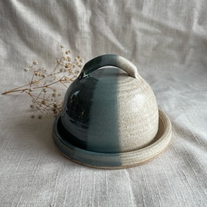 May include: A ceramic butter dish with a domed lid and a matching base. The lid is split into two vertical sections, one dark teal and the other a speckled beige. A curved handle sits on top. The base is a shallow, circular dish.