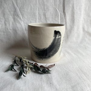 May include: A white ceramic planter with a black brushstroke design. The planter is sitting on a white fabric surface.