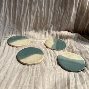 May include: Four ceramic coasters with a two-tone design. Each coaster is round, with a light beige half and a teal blue half. The coasters are arranged on a wrinkled, light pink fabric.