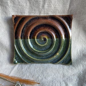 May include: Square ceramic soap dish with a spiral design. The dish has a colour gradient, with brown and black at the top, transitioning to green at the bottom. The spiral is centred, creating a unique, artistic look.