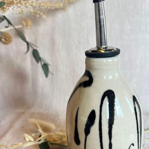 K&ouml;nnte beinhalten: Eine Keramik&ouml;lflasche mit schwarzem Ausguss und schwarz-wei&szlig;em Design. Die Flasche hat einen Ausguss aus Edelstahl und einen schwarzen Gummistopfen. Die Flasche ist mit schwarzen Pinselstrichen verziert.