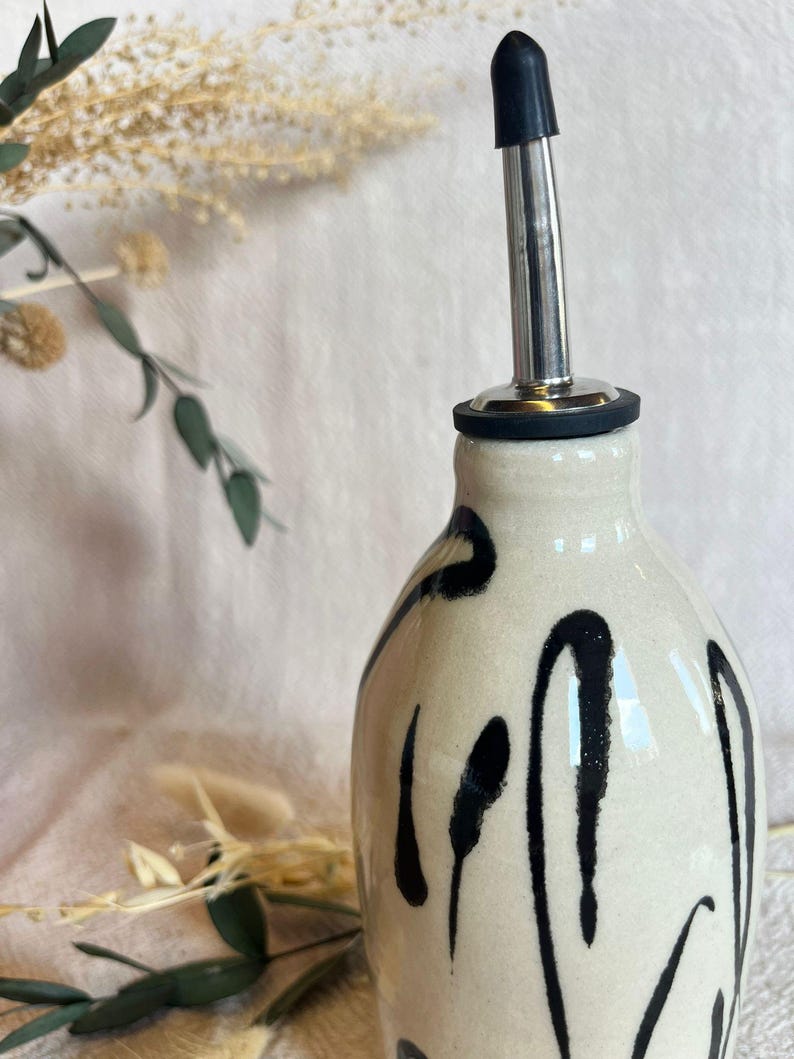 K&ouml;nnte beinhalten: Eine Keramik&ouml;lflasche mit schwarzem Ausguss und schwarz-wei&szlig;em Design. Die Flasche hat einen Ausguss aus Edelstahl und einen schwarzen Gummistopfen. Die Flasche ist mit schwarzen Pinselstrichen verziert.