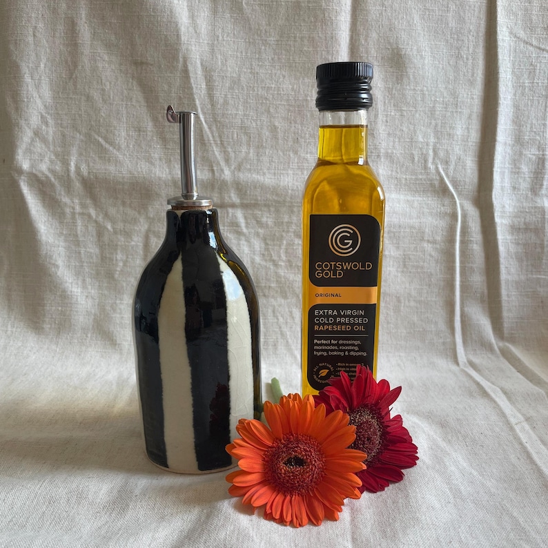 K&ouml;nnte beinhalten: Ein schwarz-wei&szlig; gestreifter &Ouml;lspender aus Keramik mit Edelstahlausguss, neben einer Flasche Cotswold Gold Raps&ouml;l. Zwei orangefarbene und rote Gerbera-G&auml;nsebl&uuml;mchen befinden sich vor den Flaschen. Die &Ouml;lflasche hat einen schwarzen Deckel.