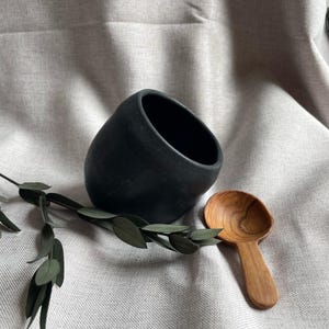 May include: A black ceramic bowl with a slanted opening, alongside a small wooden spoon. A sprig of green leaves rests on the neutral-toned fabric. The bowl and spoon are likely for kitchen or dining use.