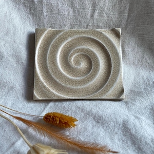 Könnte beinhalten: Eine quadratische Keramikfliese mit einem erhabenen Spiraldesign in Beige- und Weißtönen. Die Fliese hat eine strukturierte Oberfläche und ist perfekt, um jedem Raum einen Hauch von modernem Design zu verleihen.