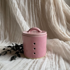 May include: A pink ceramic container with a lid and handle. The cylindrical container has three small holes and sits on a light-coloured surface. The lid has a small handle. The background is a light-coloured, textured fabric.