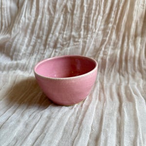 May include: A small, pink ceramic bowl with a rounded shape. The bowl has a smooth, glossy finish and a slightly lighter rim. The background is a textured, off-white fabric.