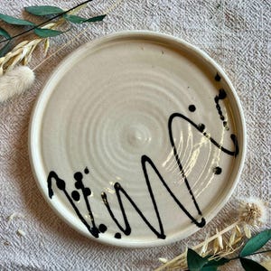 May include: A cream-colored ceramic plate with a black abstract design. The plate features a circular shape with a slightly raised rim. The black design consists of a squiggly line and several dots. The plate is a decorative item.