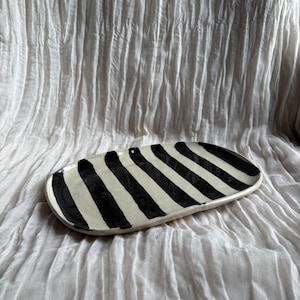 May include: An oval ceramic serving platter featuring a black and white striped design. The platter is set against a backdrop of wrinkled, off-white fabric. The stripes are consistently spaced and run the length of the platter.