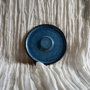 May include: A round, blue ceramic incense burner with a central raised area and a small hole. The burner has a dark rim and a textured surface. The background is a white, wrinkled fabric.
