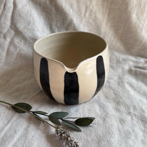 May include: A white ceramic bowl with black vertical stripes. The bowl has a rounded shape and a small spout on one side.