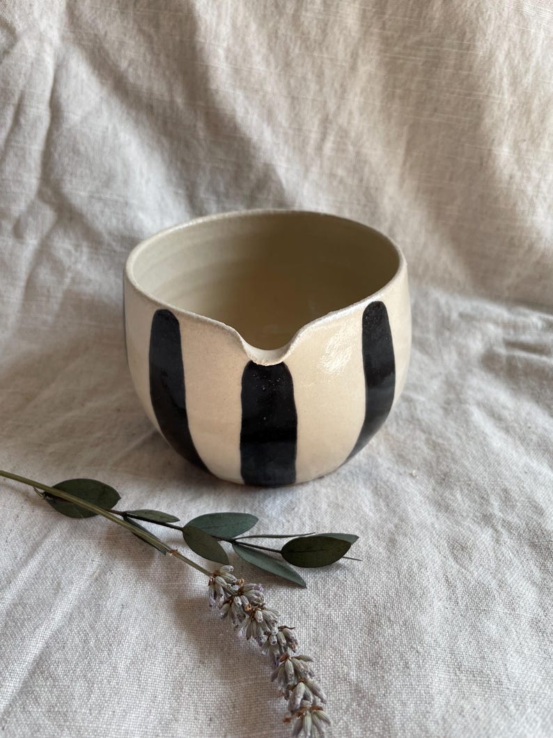 May include: A white ceramic bowl with black vertical stripes. The bowl has a rounded shape and a small spout on one side.