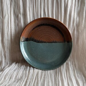 May include: A handmade ceramic plate with a two-tone design. The top half is brown with a black accent, and the bottom half is teal. The plate is round and appears to be stoneware. This plate is suitable for serving food.