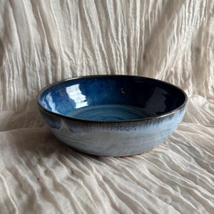 May include: A handmade ceramic bowl with a glossy, blue interior and a light blue exterior. The bowl has a dark rim and sits on a textured, off-white fabric. This unique bowl is perfect for serving food.