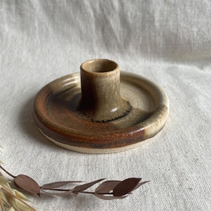May include: A brown and white ceramic candle holder with a simple design. The holder has a wide base and a narrow opening for a candle.