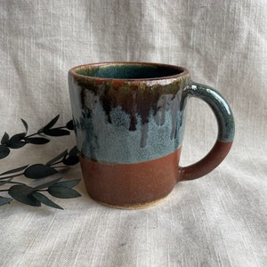 May include: A brown and blue ceramic mug with a drip glaze. The mug has a rounded body and a handle.