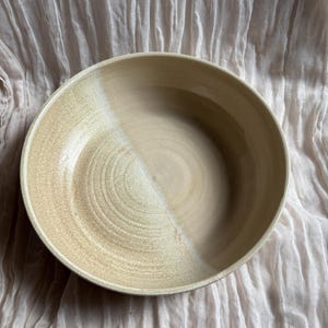 May include: A handcrafted ceramic bowl with a light beige glaze. The bowl has a wide, shallow design with a circular shape. The interior features a spiral pattern, and the bowl is set against a textured, light-coloured fabric.