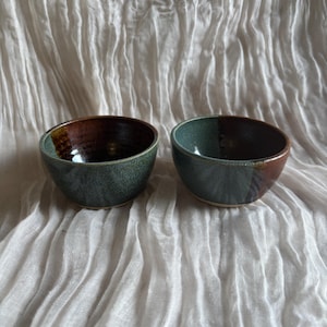 May include: Two handmade ceramic bowls. Each bowl features a unique colour scheme, combining green and brown hues. The bowls are round with a smooth, glazed finish. The bowls are set against a white, textured background.