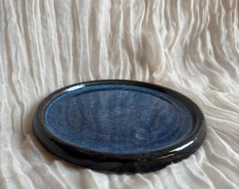 Può includere: Un piatto in ceramica fatto a mano con un centro blu scuro e un bordo nero. Il piatto ha una finitura lucida ed è di forma rotonda. Il piatto è appoggiato su una superficie bianca e strutturata.